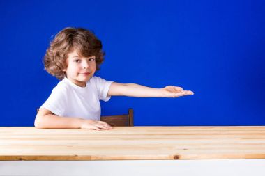 Komik kıvırcık sevimli çocuk oturan yarı-uzanmış elini ve kameraya bakarak gülümseyerek döndü. Yakın çekim. Mavi arka plan.