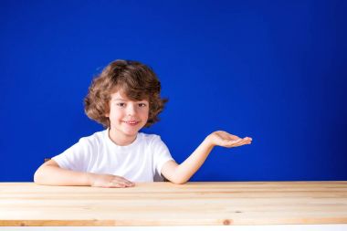 Gülümseyen ve ileri gösteren bir el kameraya bakarak mutlu kıvırcık şirin çocuk. Yakın çekim. Mavi arka plan.