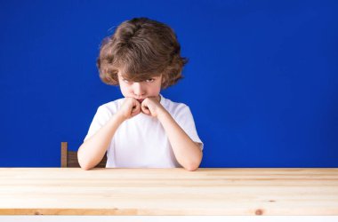 Elleri ile kafasına propped kamera bakarak düşünceli kıvırcık sevimli çocuk. Yakın çekim. Mavi arka plan.