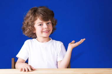 Kameraya bakarak onun sol el palm bir gülümseme ile yükselterek kıvırcık sevimli çocuk. Mavi arka plan.