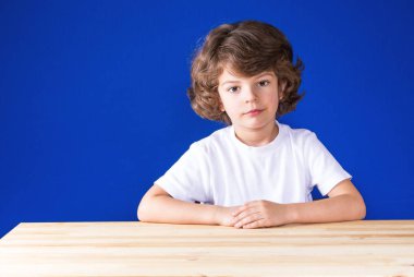 Ciddi kıvırcık şirin çocuk ahşap bir masa ve ne yazık ki oturan kameraya benziyor. Yakın çekim. Mavi arka plan.