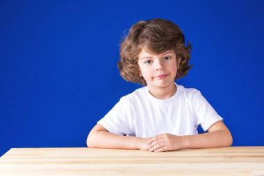 Ahşap bir masada oturuyor ve düşünceli kameraya bakarak kurnaz kıvırcık şirin çocuk. Yakın çekim. Mavi arka plan.