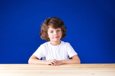 Düşünceli kıvırcık yakışıklı beyaz bir gömlek gülümseyen ve kameraya bakarak ahşap bir masaya oturur. Yakın çekim. Mavi arka plan.