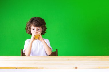 Kıvırcık sevimli çocuk kameraya bakarak masada oturan cam suyu içer. Yakın çekim. Yeşil arka plan.