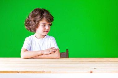 Kafasını çevirerek seyir masada kıvırcık saçlı güzel çocuk elleri katlanmış. Masaya oturur. Yakın çekim. Yeşil arka plan.