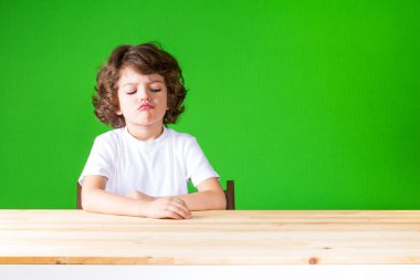 Dudaklarını güvensizlik içinde dışarı yapışmasını kıvırcık oğlan masada görünüyor. Yakın çekim. Yeşil arka plan.