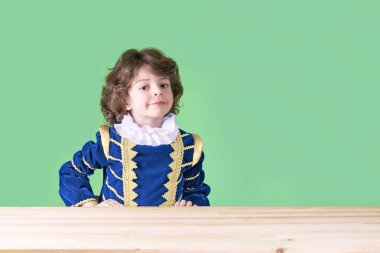 Kameraya bakarak masada elleriyle Prens giysili sevimli kıvırcık saçlı çocuk. Yakın çekim.