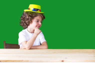 Başını sola arıyorsunuz gülümseyerek el üzerinde dinlenme bir sarı palyaço şapkalı küçük kıvırcık çocuk. Yakın çekim. Yeşil arka plan.