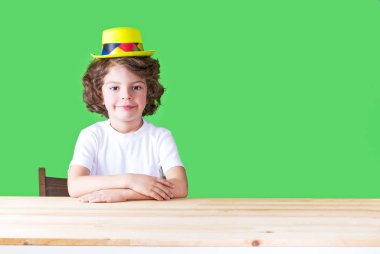 Sarı palyaço şapkalı küçük kıvırcık çocuk ellerini masanın üstünde, kameraya bakarak katlanmış. Yakın çekim. Yeşil arka plan.