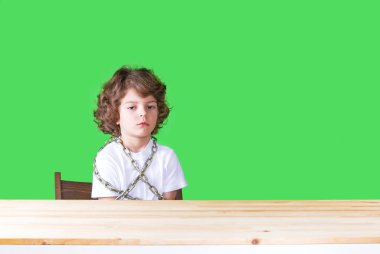 Kırgın biraz Kıvırcık kafalı çocuk Beyaz gömlekli bir sandalyeye zincirlenmiş bir masada ne yazık ki kameraya bakarak oturur. Yakın çekim. Yeşil arka plan.