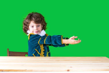 Prensin elbiseli küçük çocuk kameraya bakarak hayali bir nesne üzerinde iki el ele tutuşarak bir masada oturur. Yakın çekim. Yeşil arka plan.