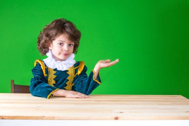 Kameraya bakarak hayali bir şey, sol elinde tutan bir masada oturan sevimli küçük prens. Yakın çekim. Yeşil arka plan.