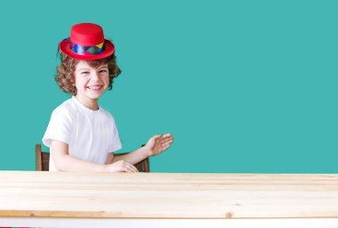 Beyaz tişört ve gülüyor, sallayarak ve kameraya bakarak bir masada oturan kırmızı şapka palyaço küçük kıvırcık çocuk. Yakın çekim. Mavi arka plan.