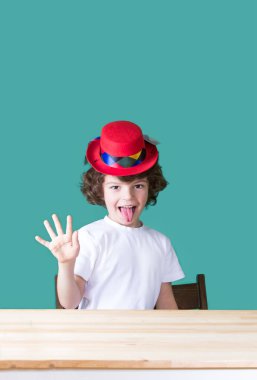 Beyaz tişört ve masada, dilini dışarı asılı, açık bir el gösterilen ve kameraya bakarak otururken koyu kırmızı şapka palyaço küçük kıvırcık çocuk. Yakın çekim. Mavi arka plan.