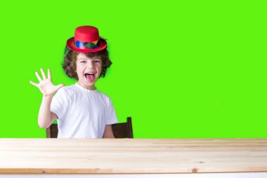 Beyaz tişört ve masada, dilini dışarı asılı, beş parmak gösterilen ve kameraya bakarak otururken koyu kırmızı şapka palyaço küçük kıvırcık çocuk. Yakın çekim. Mavi arka plan.