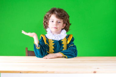 Kameraya bakarak hayali bir nesne tutarak sağ taraftan bir masada oturan sevimli küçük prens. Yakın çekim. Yeşil arka plan.