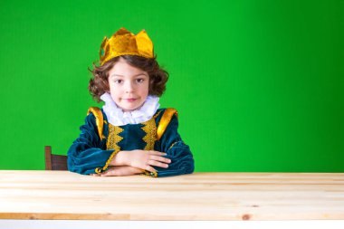 Küçük sevimli Kral gülümseyerek, kameraya bakarak masada oturuyor. Yakın çekim. Yeşil arka plan.