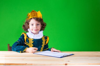 Küçük sevimli Kral bir masada oturur ve bir not defterinde yazıyor. Yakın çekim. Yeşil arka plan.