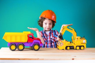 Yanındaki inşaat makinaları, inşaat kask içinde küçük kıvırcık çocuk hayali nesneleri tutar. Yakın çekim. Mavi arka plan.