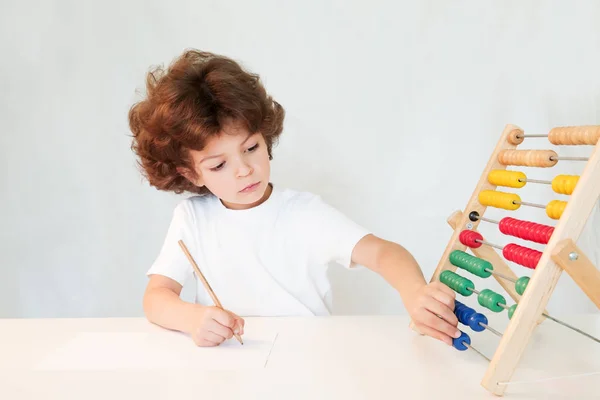 Χαριτωμένο σγουρά μαλλιά αγόρι με ένα μολύβι στο χέρι του κινεί τα οστά των βαθμολογιών. Γκρο πλαν. Γκρίζο φόντο. — Φωτογραφία Αρχείου