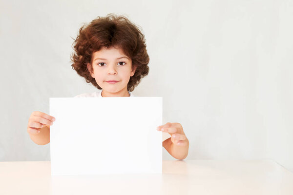 Happy cute little boy holding a blank white sheet of paper with interest and looking at the camera. Gray background.