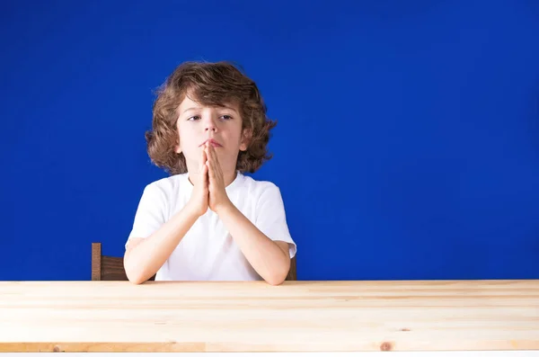 Kıvırcık sevimli çocuk ellerini dua ve üzgün dört gözle sıktı. Yakın çekim. Mavi arka plan. — Stok fotoğraf