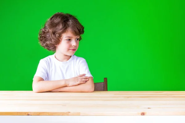 Kafasını çevirerek seyir masada kıvırcık saçlı güzel çocuk elleri katlanmış. Masaya oturur. Yakın çekim. Yeşil arka plan. — Stok fotoğraf
