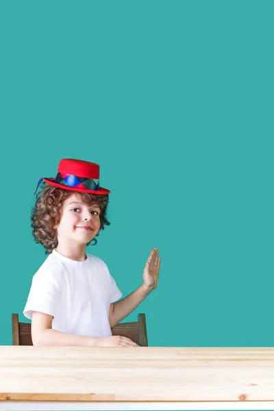 Beyaz bir gömlek ve bir masada oturan kırmızı şapka palyaço küçük kıvırcık çocuk yarı açık, kameraya bakarak palmiye ileri, elini kaldırdı. Yakın çekim. Mavi arka plan. — Stok fotoğraf