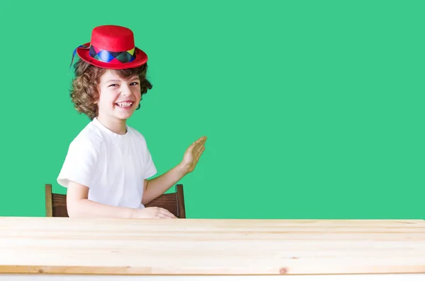 Lite lockigt pojke i en vit skjorta och en röd hatt clown skrattar, vifta med handen och tittar på kameran. Närbild. Blå bakgrund. — Stockfoto