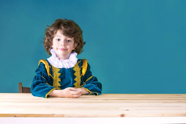 Mignon garçon aux cheveux bouclés en vêtements Prince plié les mains sur la table en regardant la caméra. Gros plan. Fond bleu . — Photo