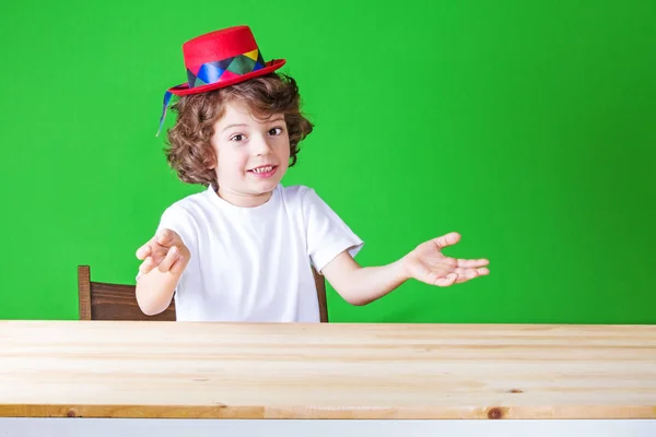 Beyaz bir gömlek ve heyecanla el hareketi koyu kırmızı şapka palyaço küçük kıvırcık çocuk diyor ve kameraya benziyor. Yakın çekim. Yeşil arka plan. — Stok fotoğraf