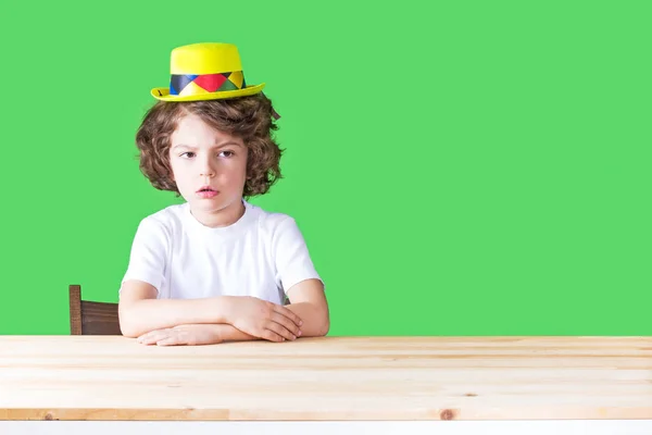 Sarı palyaço şapka ağzında küçük kıvırcık çocuk açın düşünceli kameraya bakıyor. Yakın çekim. Yeşil arka plan. — Stok fotoğraf