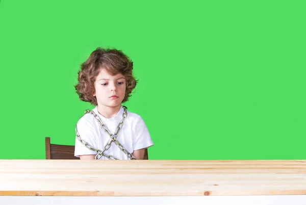 Kırgın biraz Kıvırcık kafalı çocuk Beyaz gömlekli bir sandalyeye zincirlenmiş bir masada oturur. Yakın çekim. Yeşil arka plan. — Stok fotoğraf