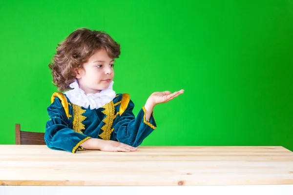 Mignon petit prince assis à une table tenant dans sa main gauche une chose imaginaire, le regardant. Gros plan. Fond vert . — Photo
