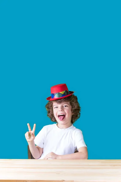 Kleiner Lockenkopf in weißem T-Shirt und rotem Hut, Clown, der lachend an einem Tisch sitzt, die Zunge herausstreckt, Geste "Sieg" zeigt und in die Kamera blickt. Nahaufnahme. blauer Hintergrund. — Stockfoto