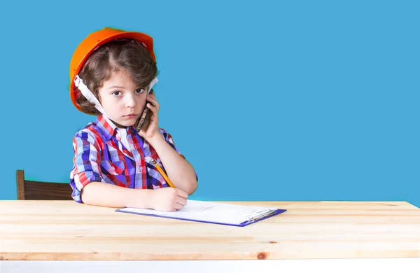 Bir telefon tutarak kask içinde genç ciddi yakışıklı foreman bir defter kalem yazdı. Yakın çekim. Mavi arka plan. — Stok fotoğraf