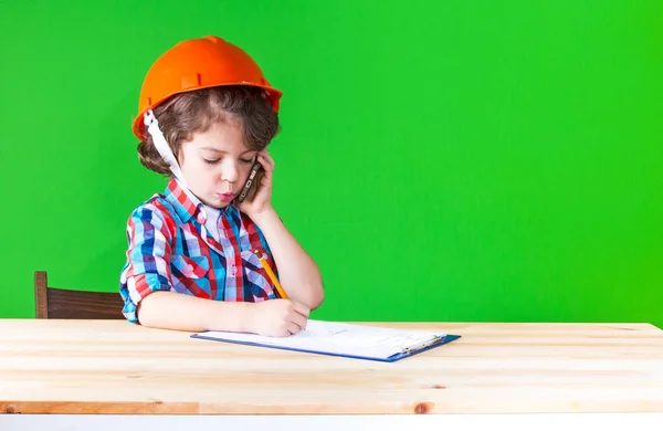 Genç sevimli baş komiser kask bir defterde yazılı onun dudak kalemi dışarı yapışmasını telefonda konuşuyor. Yakın çekim. Yeşil arka plan. — Stok fotoğraf