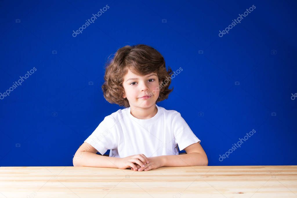 Thoughtful curly cute boy in a white shirt sits at a wooden table, smiling and looking at the camera. Close-up. Blue background.