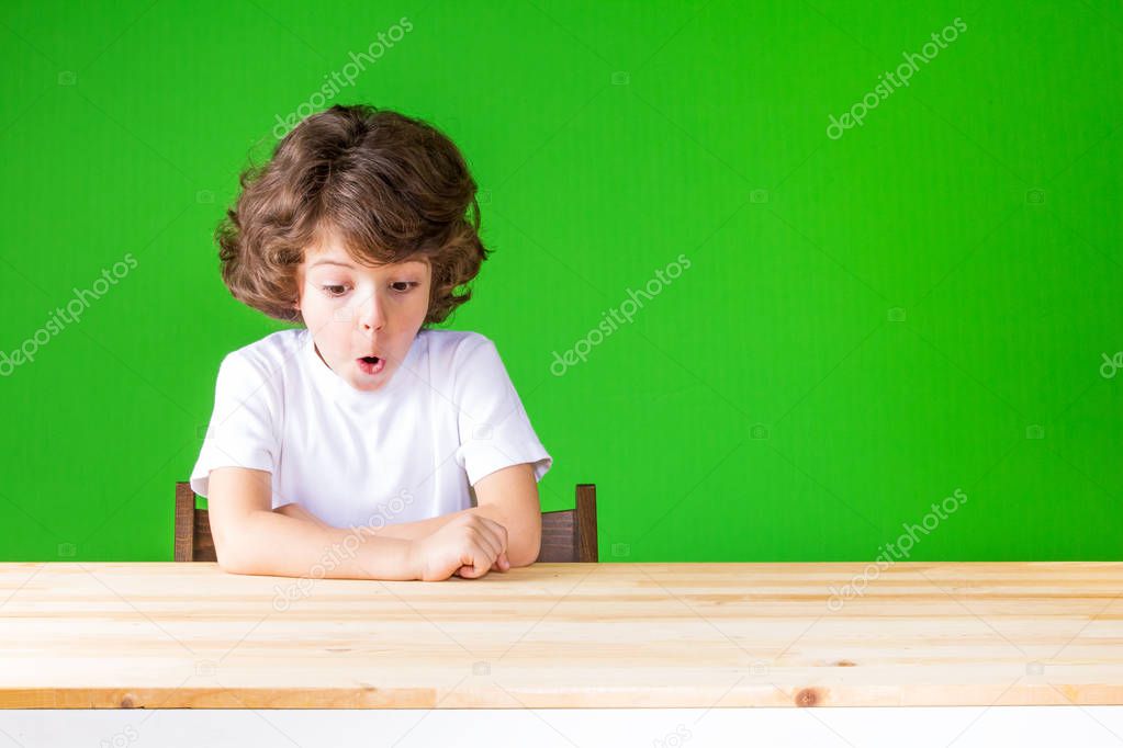 Curly resentful little boy sitting with his mouth open, looking at the table. Close-up. Green background.