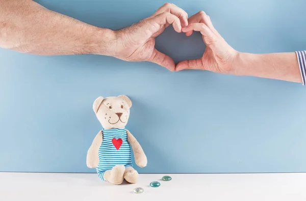 Mäns och kvinnors hand göra symbol ”hjärtat”. Därunder är en nallebjörn. Blå bakgrund. — Stockfoto