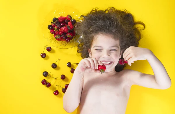 Funny and happy kid eating a berries. Kid lies on the yellow background .Dark hair, pigtails. Strawberries, cherries ripe, red. The kid laughs — Stock Photo, Image