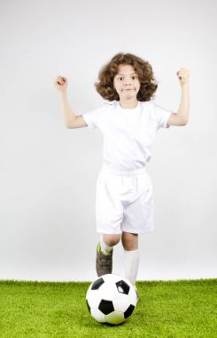 Yumruklarını zafer bir çocuk futbol forma ve şort. Gri bir arka plan üzerinde bir futbol ile mutlu çocuk.