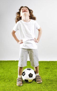 Küçük çocuk beyaz şort ve beyaz gömlek bir futbol topuyla oynuyor. Eğlenceli çocuk sesle gülüyor.