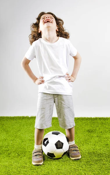 Küçük çocuk beyaz şort ve beyaz gömlek bir futbol topuyla oynuyor. Eğlenceli çocuk sesle gülüyor.
