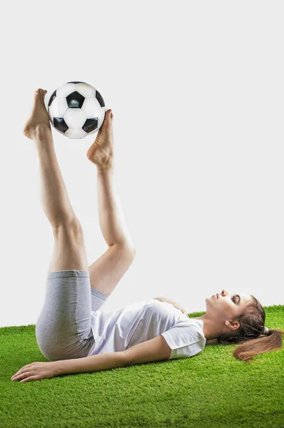 Fútbol femenino bonito. Joven mujer de cabello castaño en ropa deportiva se encuentra con una pelota de fútbol en la hierba, aislado sobre fondo gris —  Fotos de Stock