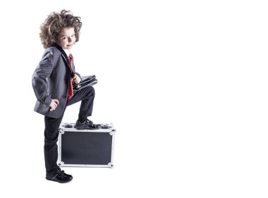 Küçük kıvırcık saçlı çocuk işinde giymiş siyah bir çanta ile ayakta uygun, çanta ve beyaz bir arka plan üzerinde izole kamera bakar. Çocuk ayağı bavula koy..