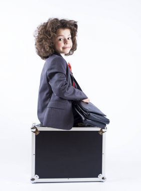 Küçük kıvırcık saçlı çocuk kameraya seyir siyah bavul üzerinde oturan bir takım elbise giymiş. Gri bir arka plan üzerinde izole.