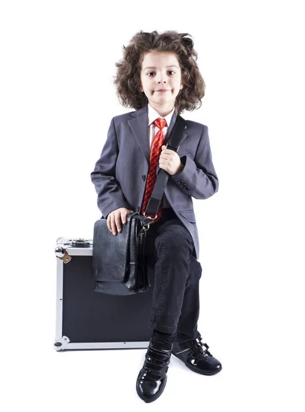 Kleiner lockiger Junge, der auf dem schwarzen Koffer sitzt und isoliert auf weißem Hintergrund in die Kamera schaut — Stockfoto