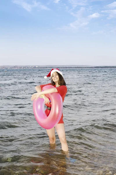 Різдвяний портрет красива жінка в червоному капелюсі Санта з колом надувний робить повітряний поцілунок над морським тлом — стокове фото