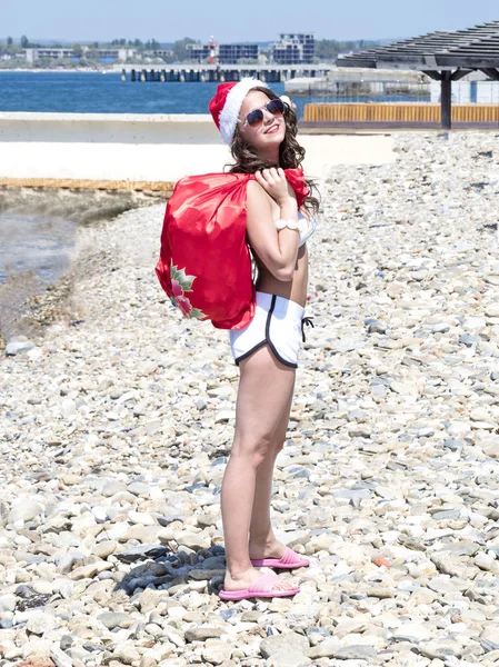 クリスマスと新年のお祝い。海のビーチを歩いてプレゼントの袋を持つ女性。サンタ衣装を身に着けている水着の女の子。冬の休日の概念 — ストック写真